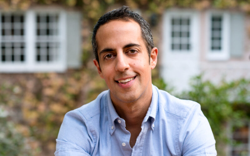 Photo of Abdi Nazemian. He has short dark hair and is smiling. He's wearing a light blue shirt and there is a slightly blurry house with ivy in the background.