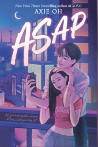 A young woman with long hair is leaning back against a young man. She's wearing light jeans and a crop top that says ASAP. He has a hand and his chin leaning on the top of her head and her hands are lifted and resting on his arms. He is holding a phone out in front of them. They both have slight smiles and it seems they are on a roof and it is early evening. 