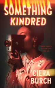 Book cover with title something kindred in pale glowing letters. The background is dark and a young Black woman is holding an old camera up to her eye. Her hair is up and she is wearing a pink floral print dress. Her face is obscured by the camera.