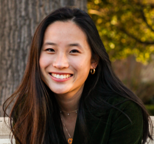 Headshot of Christina. She has long, dark, fairly straight hair. She is leaning foward and smiling. There are trees behind her. 