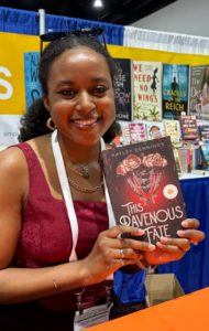 A smiling Black woman is holding up the book This Ravenous Fate. She is wearing a dark red tank dress. The book cover is also in reds with some white and dark gray accents. There are roses that are white with red edges along with clear beads on the cover.