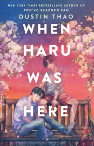 Illustrated cover of When Haru Was Here. A teenage boy sits sideways on a wooden park bench, with his head resting on the thigh of another teenage boy who is sitting on the back of the bench. That boy looks like is stroking the first boy's hair, but he is also semi-translucent. They are surrounded by blossoms, and petals are floating off in the wind. The sky makes it look like it is either approaching sunrise or sunset.
