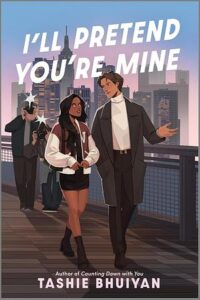 Book cover of I'll Pretend You're Mine. There is a young couple on a walkway with multiple photographers following them. She has long wavy dark hair and brown skin and is wearing a miniskirt books and a jacket. He has light brown skin, short brown hair and is wearing a white turtle neck and a long coat.