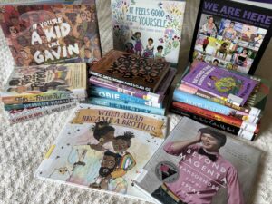 17 books on a blanket. Three are standing up If You're a kid like Gavin, It Feels Good to Be Yourself, and We Are Here. Then there are also three stacks of books and two flat in front of the stacks. When Aidan Became a Brother and Beyond Magenta. In the stack on the left is This is our rainbow, disability visibility, and Glimmer of Hope. In the middle stack is Women, Race and Class, Pet, Obie is Man Enough, and He/She/They. The stack on the right has Beyond the Gender Binary, Red to the Bone, All Boys Aren't Blue, How We Fight for our lives, and This Bridge Called My Back.