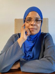 A light skinned Black woman is wearing wire rimmed glasses and a cobalt blue headscarf covers her hair and the front portion of her shirt. She is resting one hand at her fae. She has a gray long sleeved shirt or dress on. She has a slight smile. 