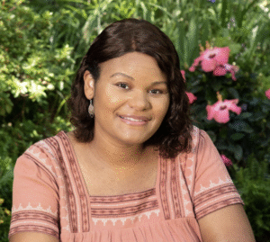 Photo of Pamela N. Harris. A smiling Black woman with shoulder length wavy hair is sitting outside with greenery and some fushia flowers behind her. She has on a peachy short sleeved blouse. 