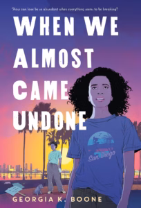 Book cover of When We Almost Came Undone. There is a young woman with shoulder length dark curly hair is looking pensively up to the sky. She is wearing a San Diego t-shirt. In the background is a cityscape with a setting sun. There is also another person standing just a little behind here with a dog on a leash. That person has short dark hair and brown skin. They are wearing a t-shirt and sweatpants. There are a few signs lying on the ground near the dog. 