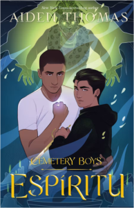 Illustrated cover for Cemetery Boys: Espíritu. Two Latine teenagers hold each other defensively as they look at the viewer. One is holding a glowing pendant necklace and the other has a gold dagger. There are wisps of smoke across the cover, and the teenagers are encircled by low-burning candles. Someone in what looks like a leopard skin is reaching out toward them as the person looms overhead.